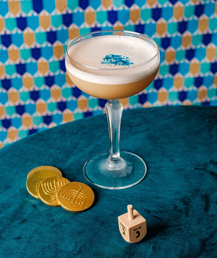 A pale pink cocktail with foam and blue sprinkle garnish sits on a blue surface, surrounded by Hanukkah gelt and a dreidel.