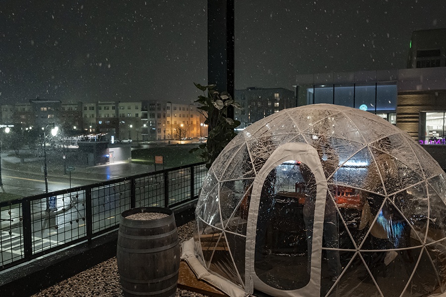 A see-through dining igloo sits atop a restaurant rooftop with city skyline views as snow falls.