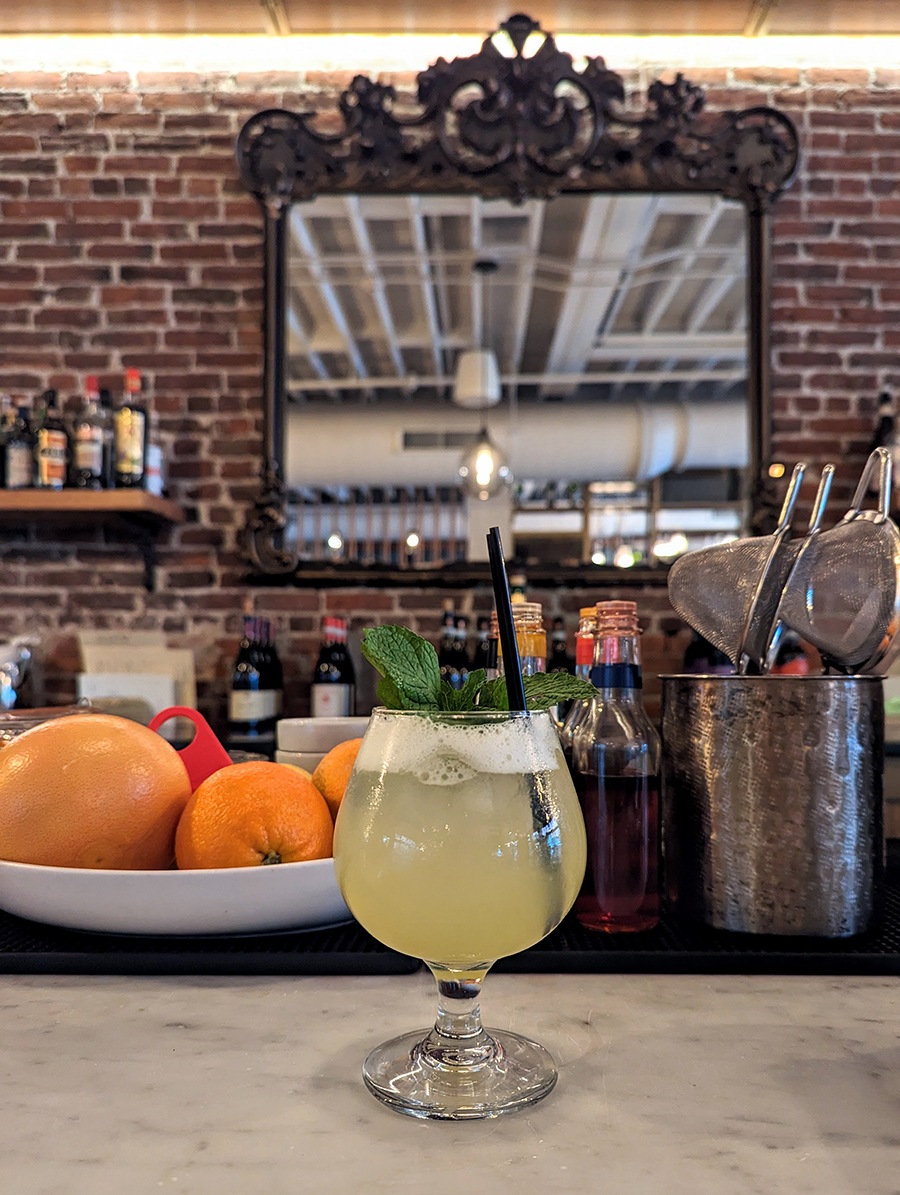 A yellow cocktail sits on a white marble bar in front of an elaborate framed mirror.