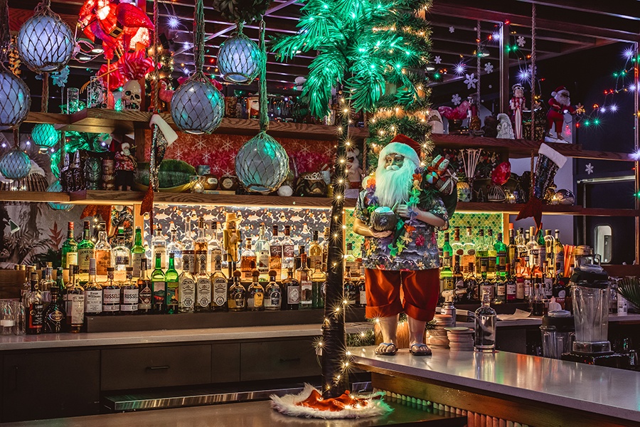 A bar is decorated with Christmas lights, a light-up palm tree, and a giant Santa figure who looks dressed for a beach and holds a tiki-style cocktail.