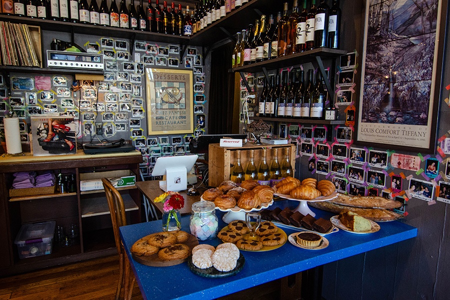 A blue table covered with baked goods sits in front of a cafe register. The walls of the space are decorated with polaroids and shelves of wine bottles.