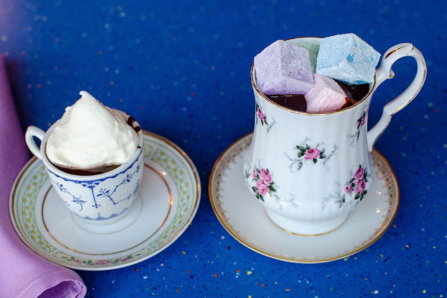 A small mug and a large mug are each filled with very thick drinking chocolate. One is topped with a dollop of whipped cream, the other with large marshmallows in a variety of pastel colors.