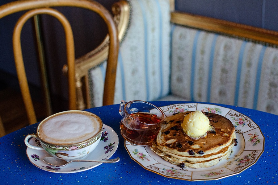 A stack of pancakes sits on a floral plate on a bright blue table, topped with butter, with a cappuccino on the side.