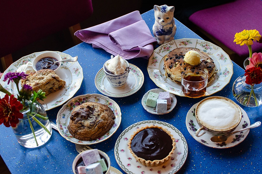 A spread of baked goods and hot drinks sit on a bright blue table with colorful flowers and a white and blue china cat.