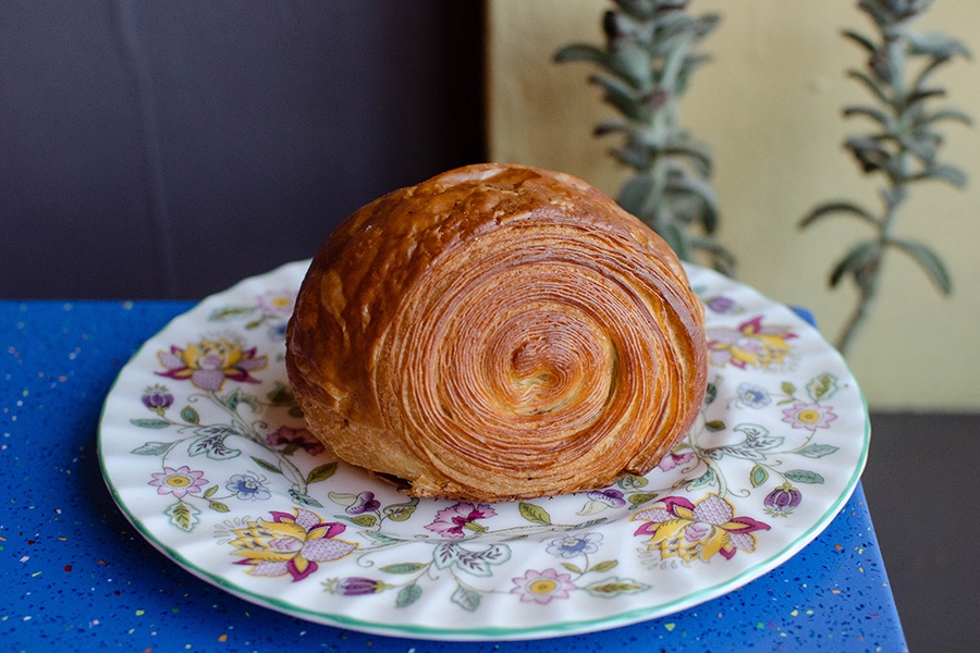 A croissant-like pastry with many layers sits on a floral plate on a bright blue table.