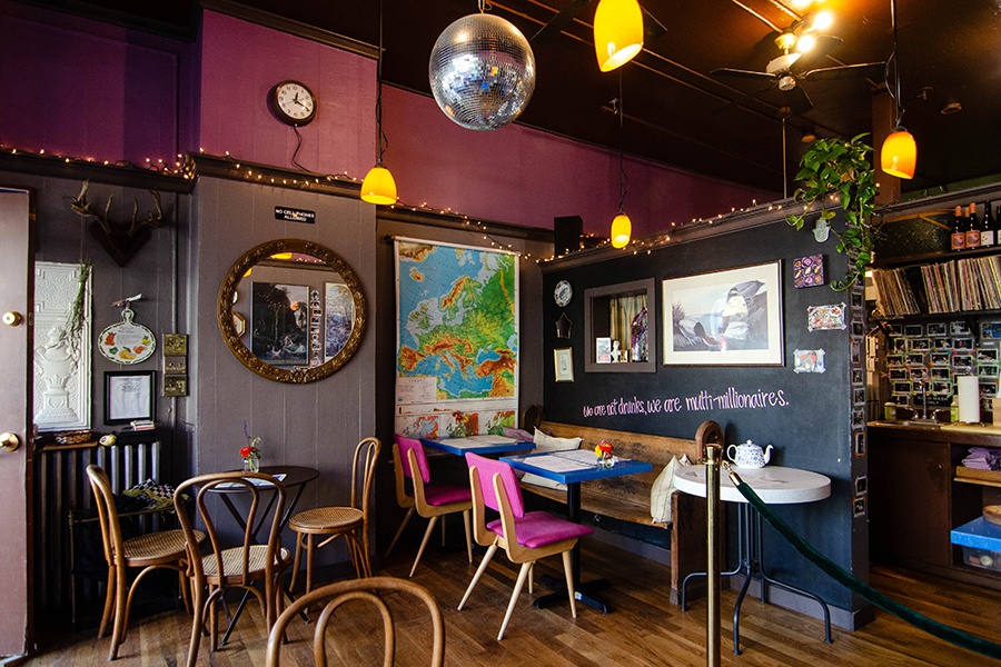 Interior of a cafe, featuring a disco ball, pink accents, a map of Europe, a big mirror with a decorative brass frame, string lights, and a quote painted in pink on a black wall: "We are not drunks, we are multi-millionaires."