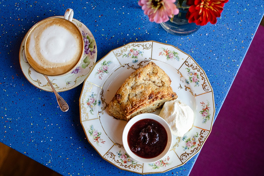 Overhead view of a cappuccino and a scone with a dollop of whipped cream and a small bowl of red jam.