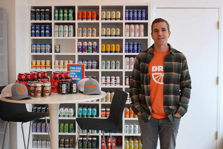 A man in flannel over an orange shirt stands in front of shelves full of cans on nonalcoholic drinks.