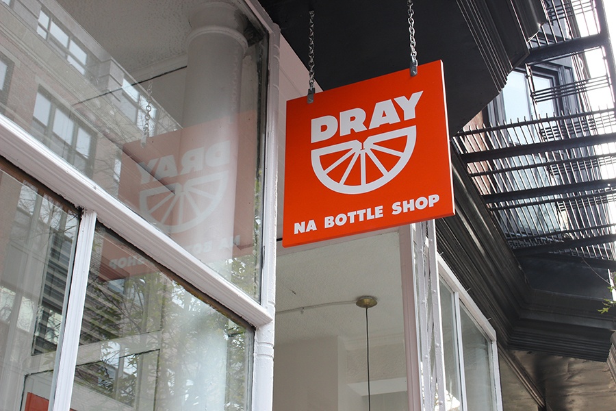 An orange, square-shaped sign that says Dray NA Bottle Shop hangs outside a city window.
