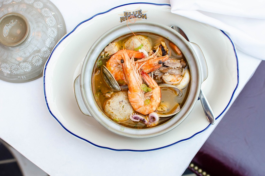 Overhead shot of a big bowl of soup with head-on shrimp, clams, matzoh balls, and more.