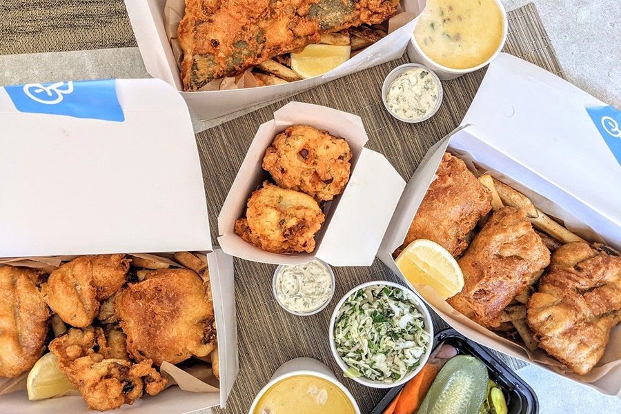 Overhead view of white paper takeout containers of fish and chips, crab cakes, chowder, and sides.