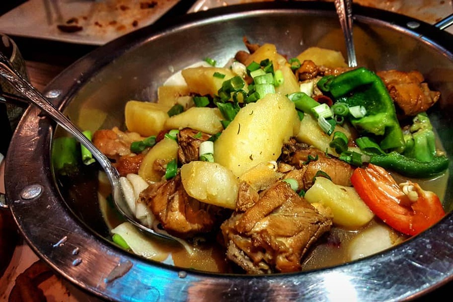A silver bowl is full of chicken chunks, potatoes, peppers, and thick noodles.