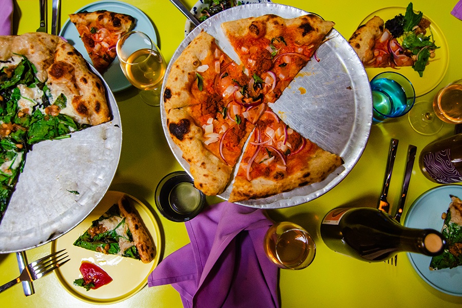 Overhead view of two pizzas with some slices taken out and spread across a bright yellow-green table with glasses of wine.