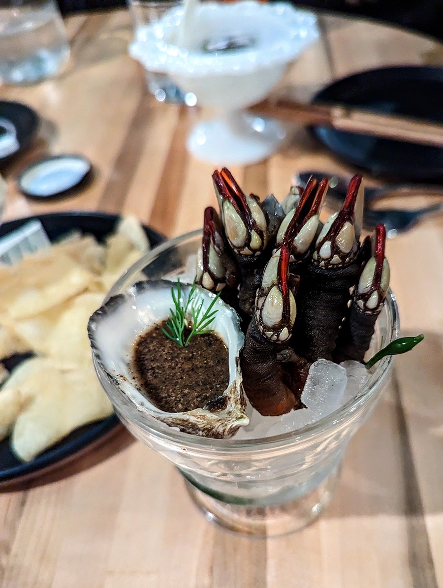 Barnacles that look like dinosaur claws come out of ice in a glass, with a small oyster shell full of dipping sauce.