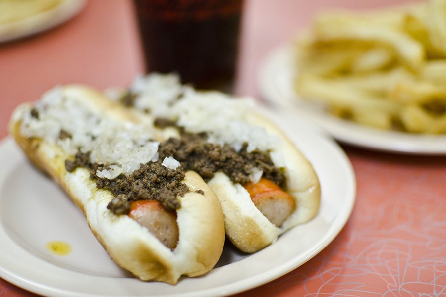 Two hot dogs on a white plate are smothered in a ground meat sauce and chopped white onion.