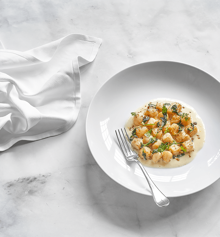 An elegant white bowl of gnocchi is on a white marble tabletop.