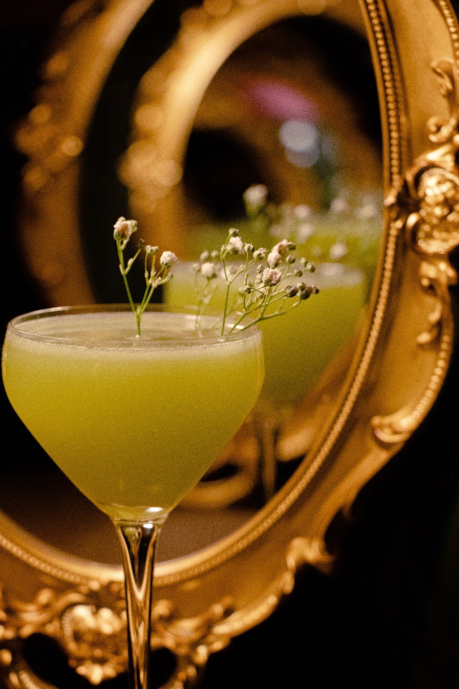 A green-yellow cocktail is garnished with sprigs of baby's breath. and reflected in a brass-framed mirror.