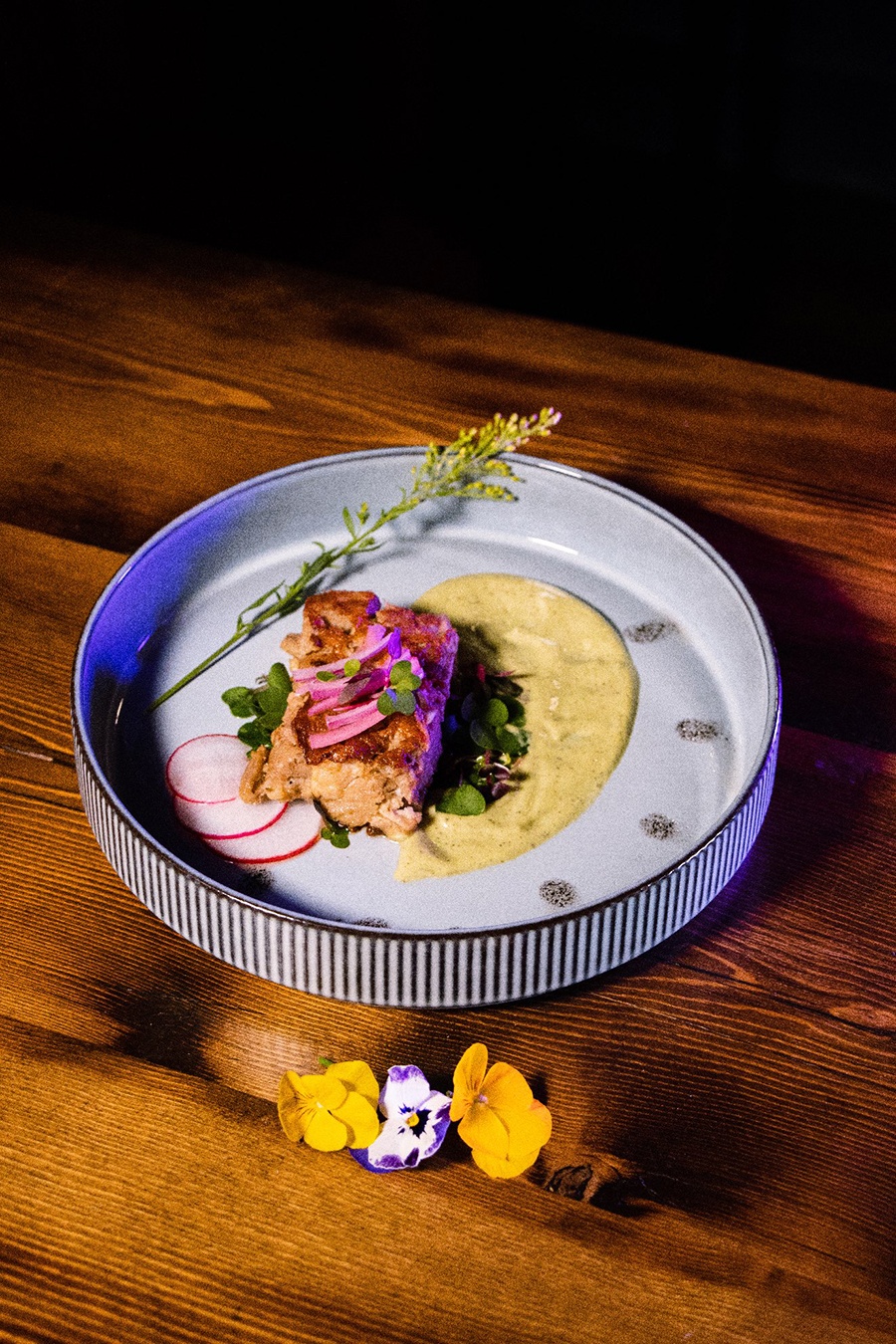 A square of pork sits on a greenish cream sauce with red onions, sliced radishes, and other garnishes.