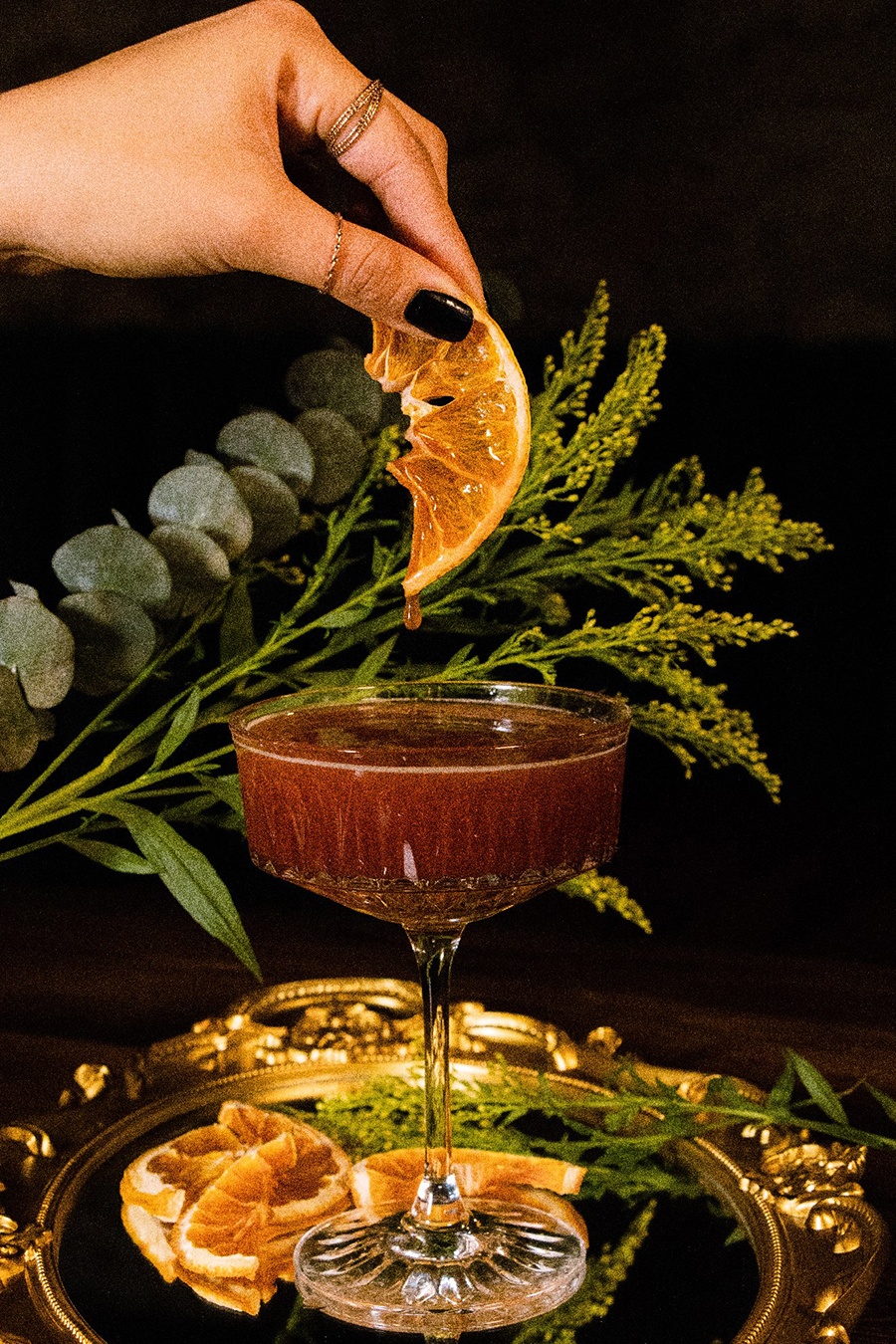 A hand squeezes juice from an orange slice into a pink cocktail.