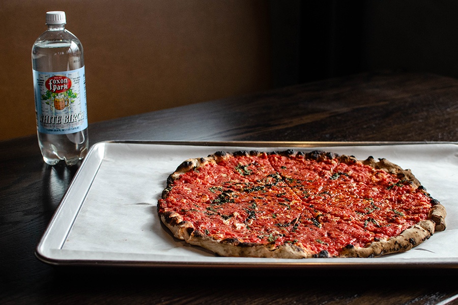 A thin-crust pizza with a charred crust sits on a metal tray on a wooden table, with a bottle of clear soda in the background.