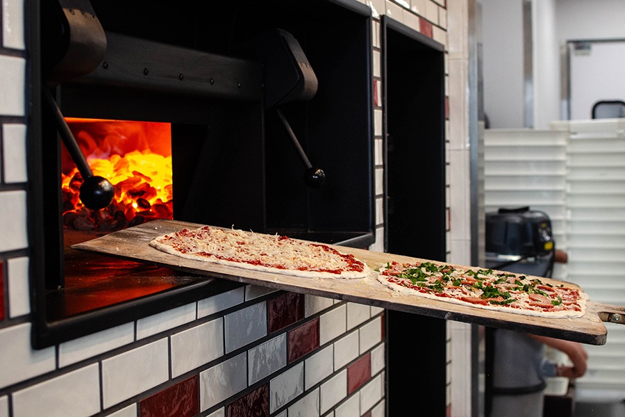 Two thin-crust pizzas are going into a fiery oven atop a large wooden pizza peel.