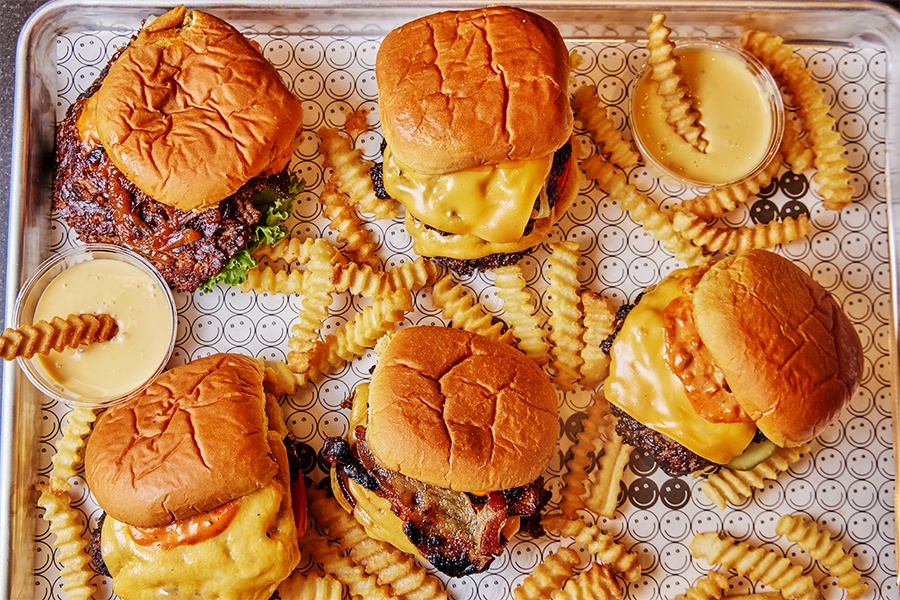 Overhead view of a tray of smashburgers and crinkle-cut fries on tissue paper covered with smiley faces.