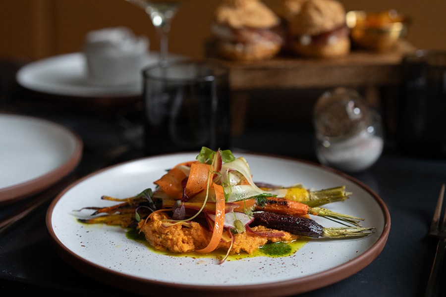 Roasted, multi-colored carrots are plated elegantly over a thick orange hummus, with vegetable ribbon garnish.