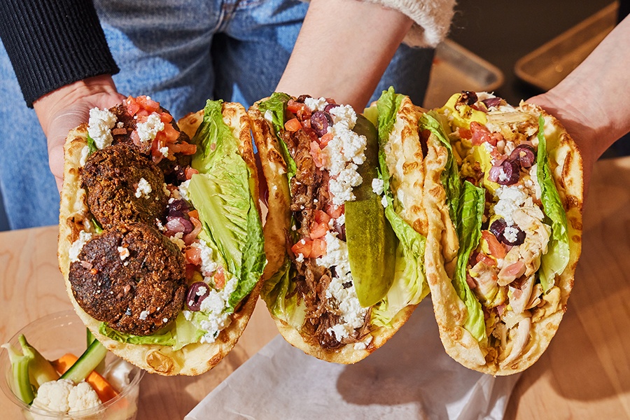 Hands hold three pitas stuffed with falafel, feta, lettuce, and other fillings.