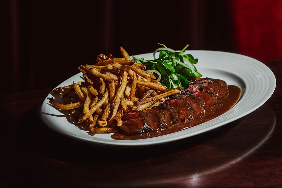 Sliced steak in a brown sauce sits on a plate with fries and greens on a dark wooden table.