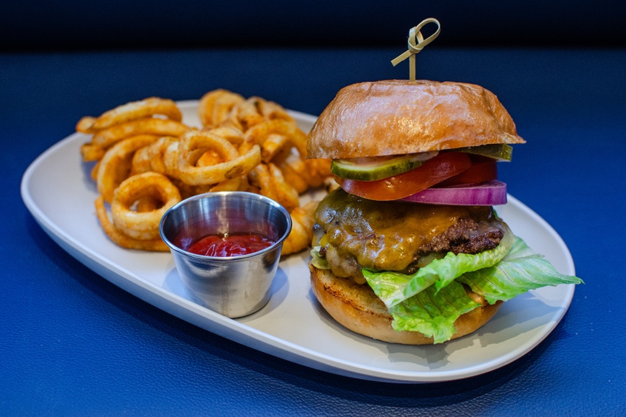 A burger, photographed on a blue background with a side of curly fries, features two smashed patties.