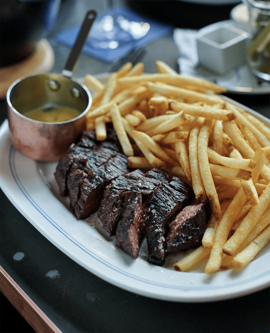 Thickly sliced steak sits on a plate with lots of fries and a small silver cup of pan sauce.