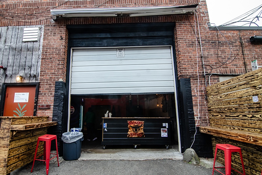 A small takeout counter is visible beyond a half-opened garage door in a brick building, with wooden counters and red stools out front.