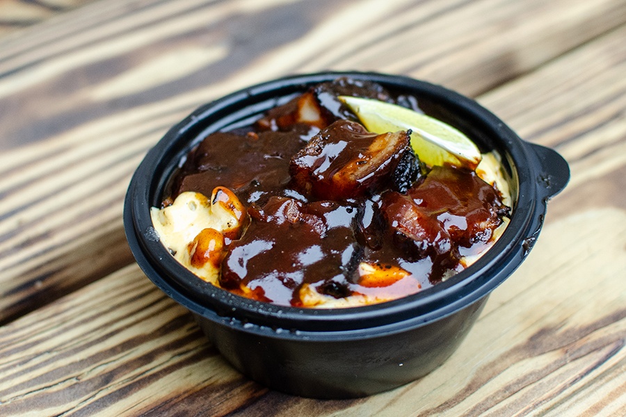 A black plastic takeout bowl sits on a wooden surface and is full of barbecue sauce-covered chunks of pork belly and a lime wedge.