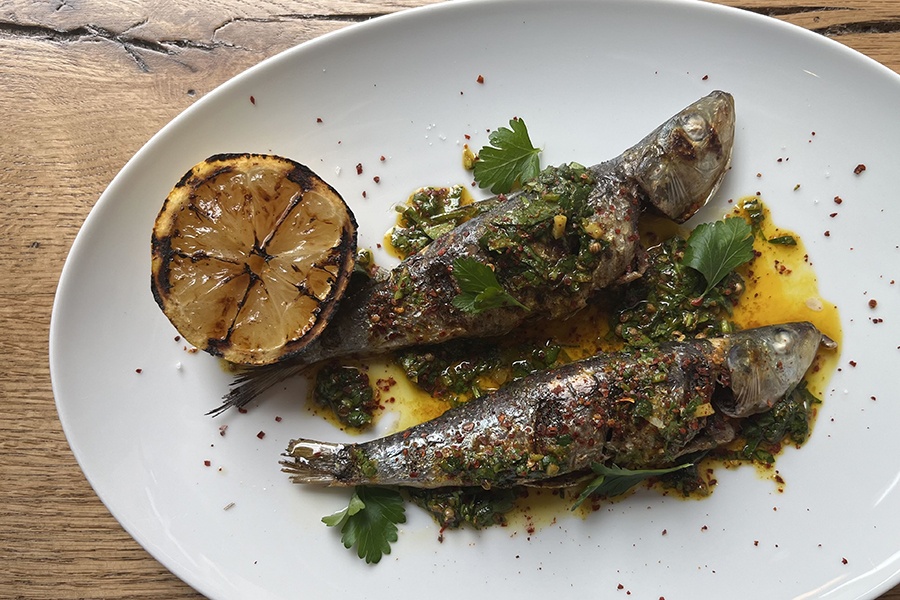 Two grilled whole fish sit in an oily, herby sauce on a white plate, accompanied by a charred lemon half.