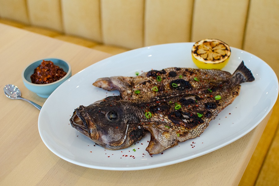 A whole fish is topped with chili flakes and sliced scallions, served with a grilled lemon half and a red condiment.