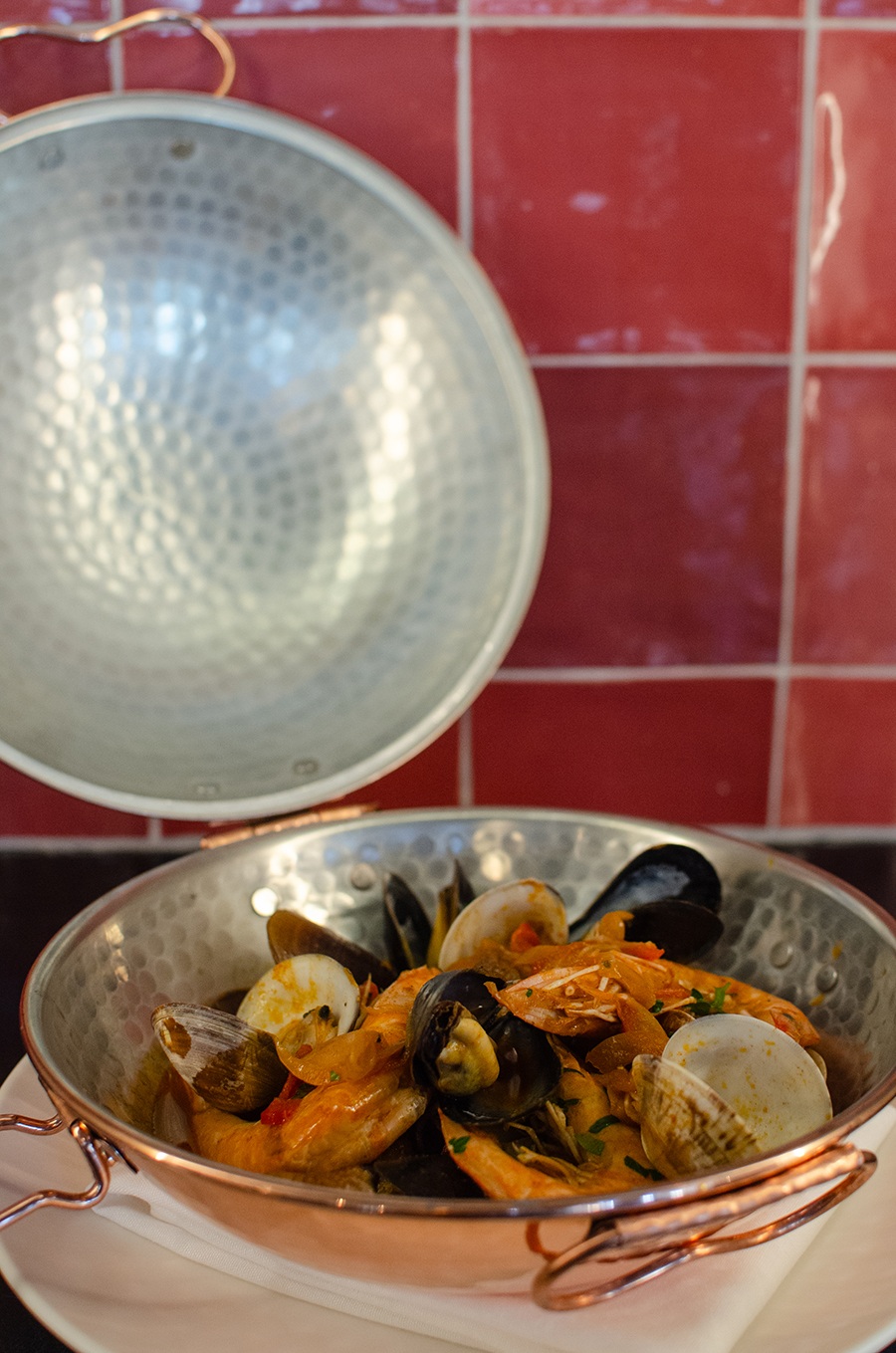 Clams, mussels, shrimp, and broth sit in a big copper pot in front of a red tiled wall.