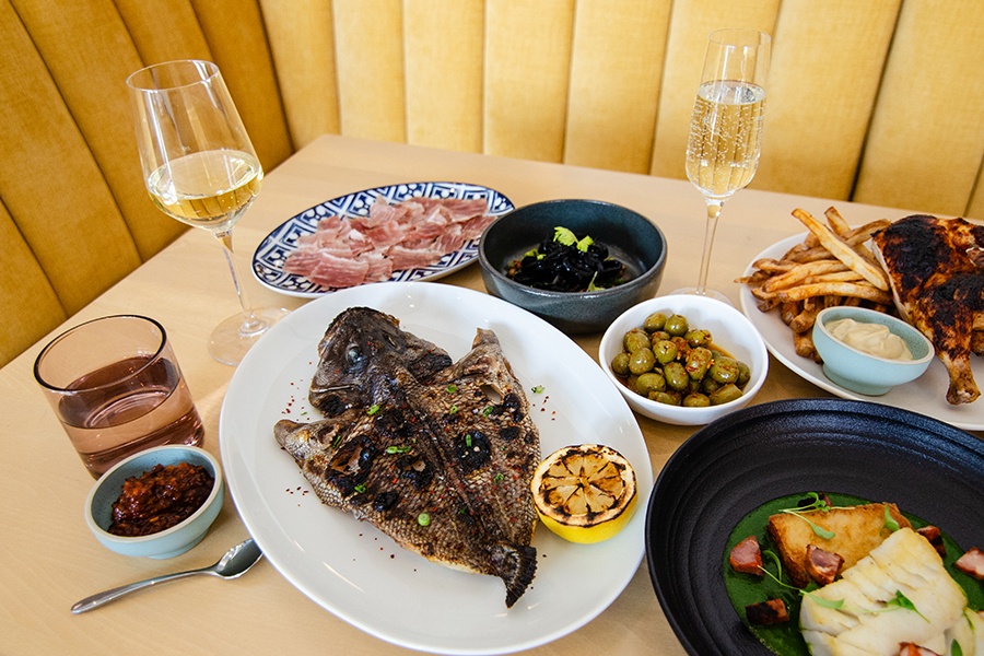 A spread of Portuguese-inspired dishes sit on a light wooden table next to a pale yellow banquette.