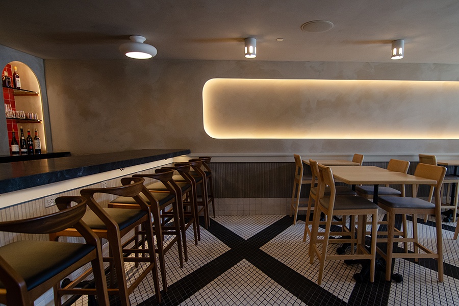 An empty restaurant space features a tiled black-and-white floor, limewashed walls, and curved-back seats at a dark marble bar.