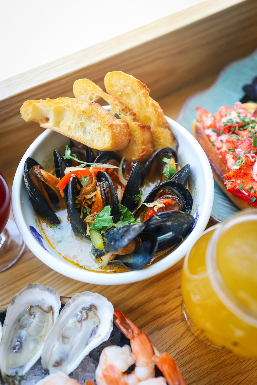 A bowl of mussels in broth, accompanied by bread, sits on a counter with other seafood dishes and glasses of beer.