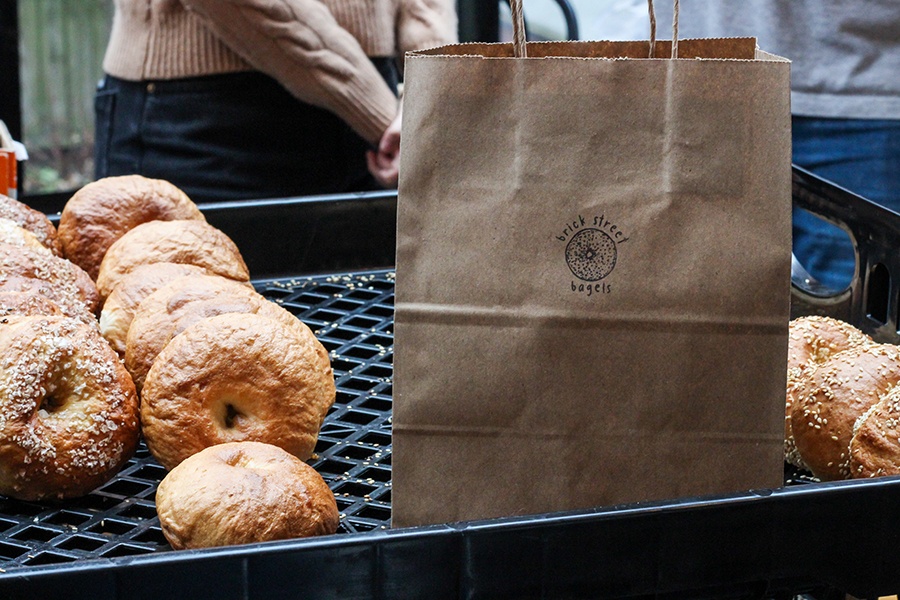 A brown bag with a stamp that says Brick Street Bagels sits in a bin alongside a few rows of bagels.