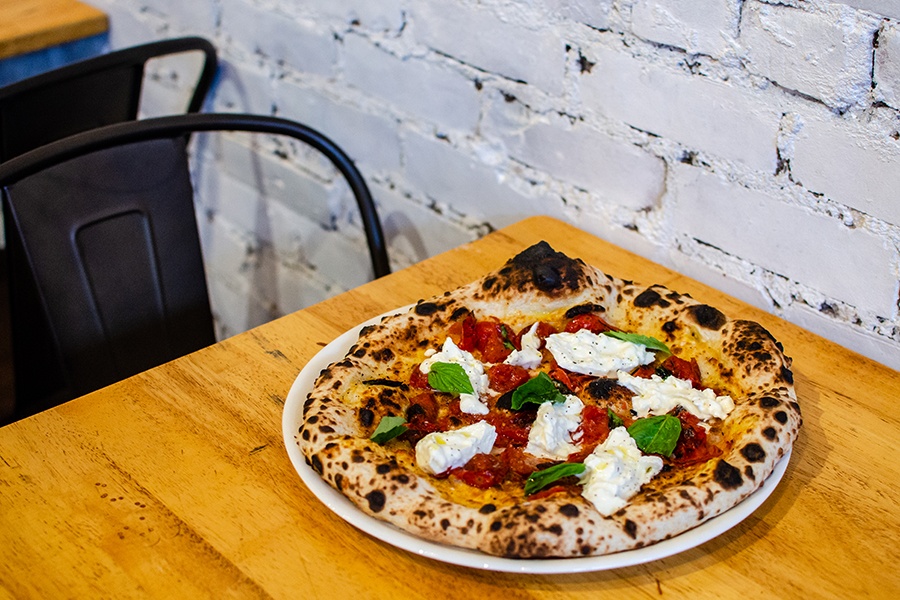 A pizza topped with chunks of burrata and roasted tomatoes sits on a wooden table in front of white brick.