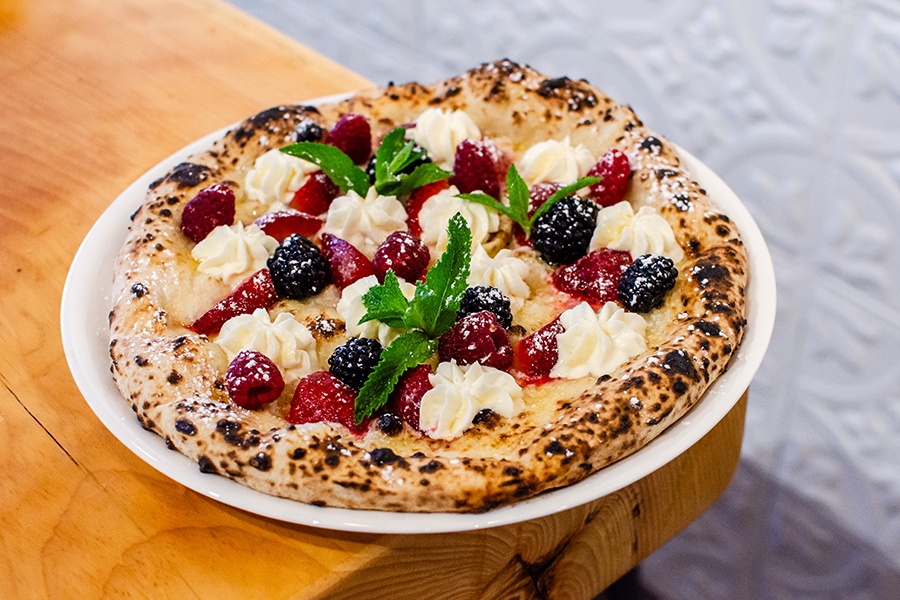 A dessert pizza with leopard-spotted crust is dotted with dollops of cream, fresh berries, mint leaves, and powdered sugar.