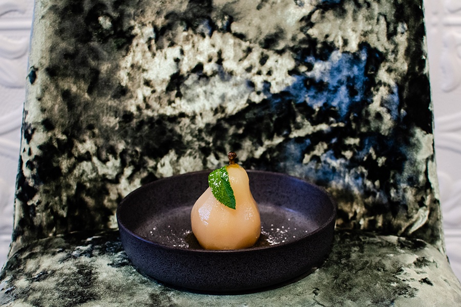 A poached pear, garnished with a mint leaf, sits on a dark plate on a green crushed velvet chair.