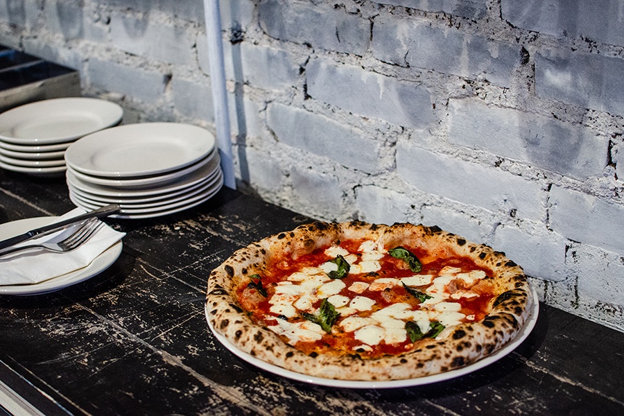 A margherita pizza with a leopard-spotted crust sits on a rustic dark wood countertop in front of a white brick wall.