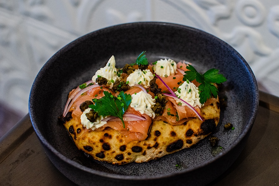 A round, thick piece of pizza dough is topped with pieces of smoked salmon and dollops of cream cheese.