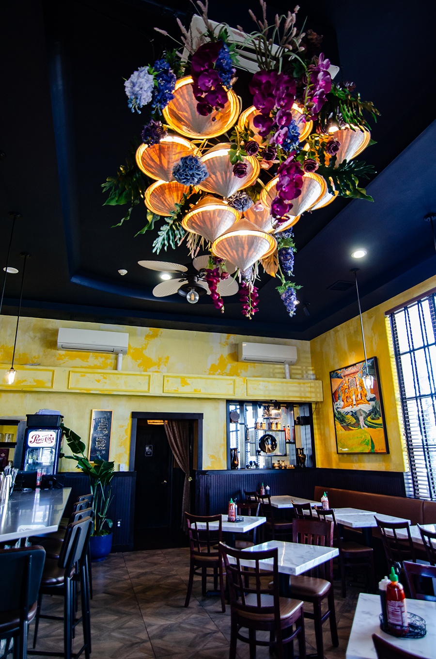 A restaurant dining room features yellow walls, a dark wooden floor, and an intricate light fixture of flowers and Vietnamese hats.