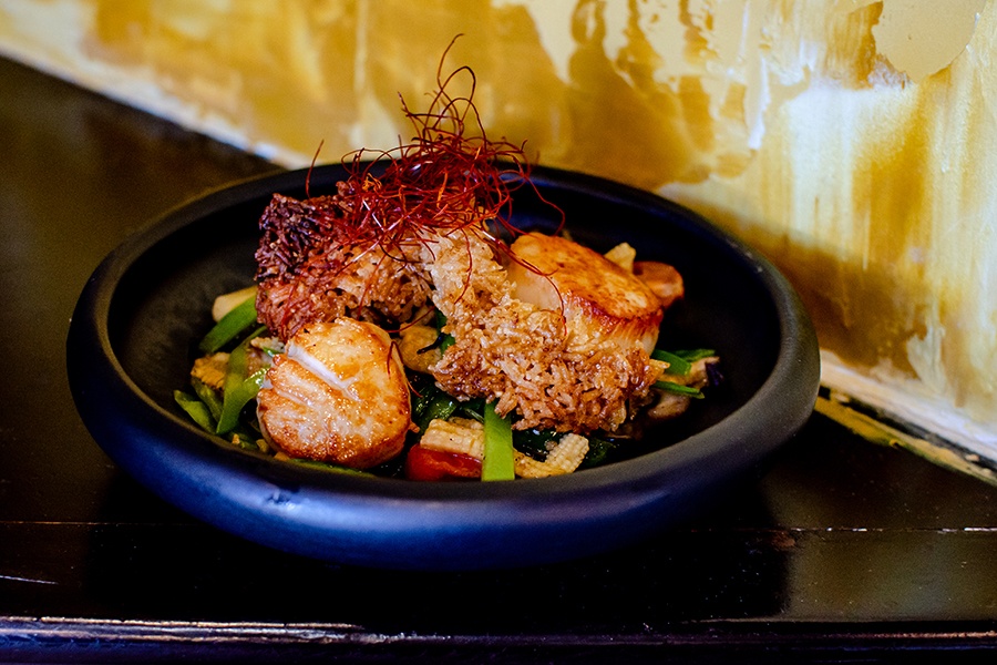 Seared scallops, crispy rice, and vegetables sit in a black bowl.