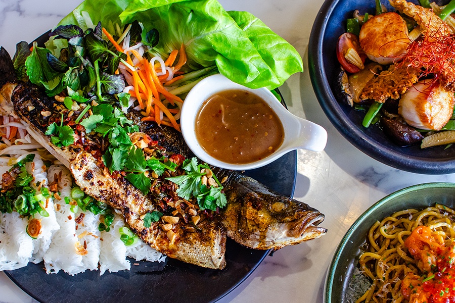 Overhead view of an herb-covered whole roasted fish with vermicelli next to a plate of seared scallops and a plate of crawfish and noodles.