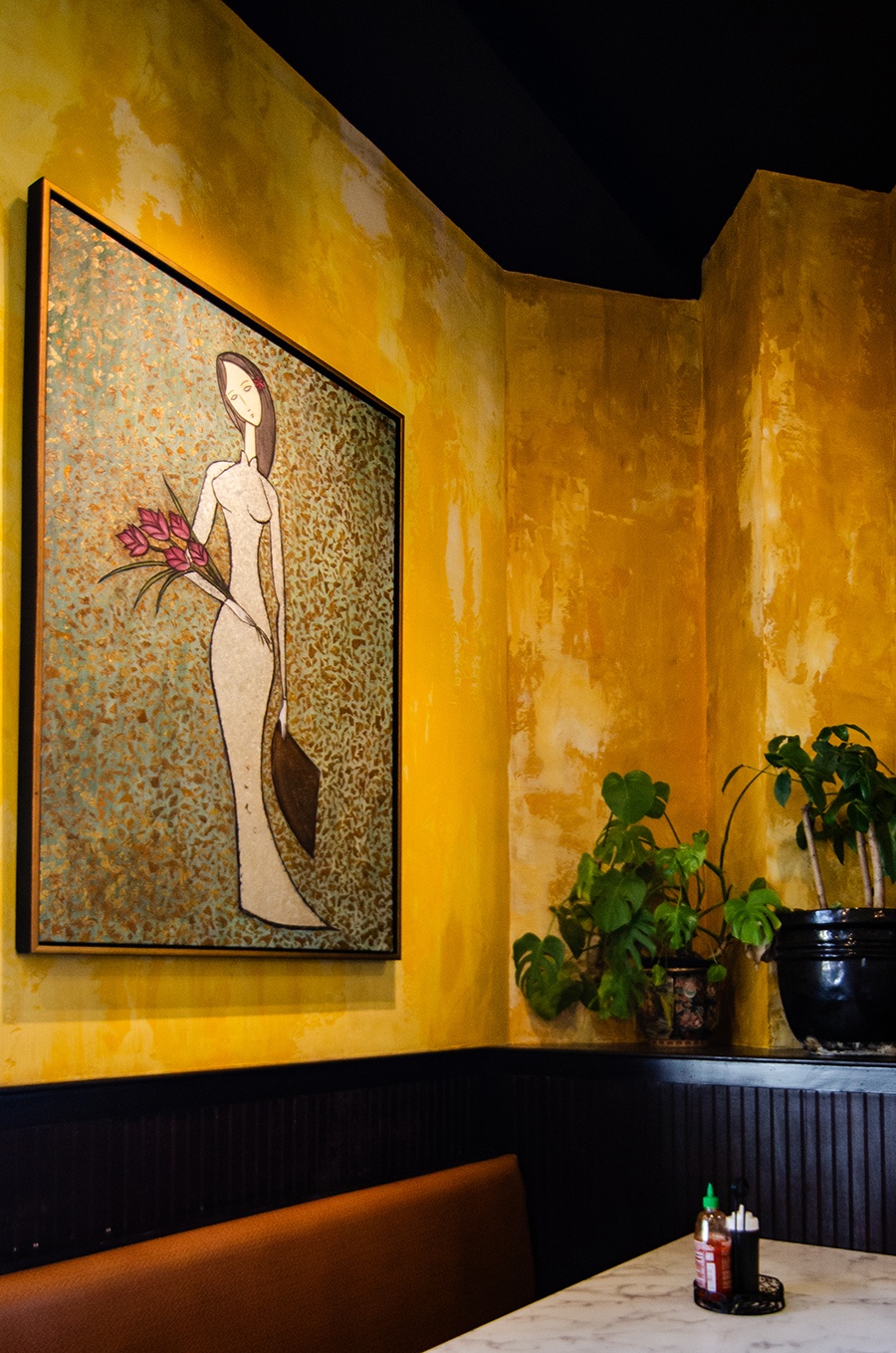 A corner of a restaurant with yellow walls features a painting of a woman holding flowers.