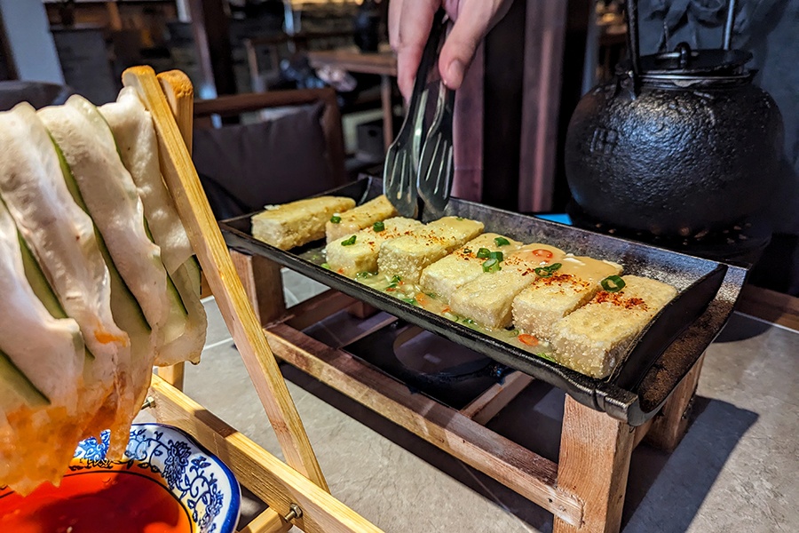 Crispy rectangles of tofu are cooked in a rectangular cast-iron pan at a restaurant table, with a dish of thinly sliced pork belly hanging on a wooden rack visible in the foreground.